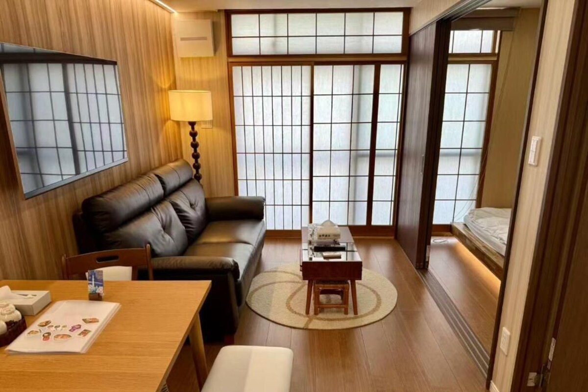 A traditional Japanese-style room with tatami flooring, a low table with a lamp, a sofa, and a sliding door leading to a sleeping area.