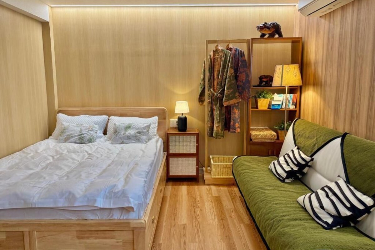 A bedroom with a bed, a green sofa, a nightstand, a lamp, a wardrobe, and a shelf with books and decorative items.