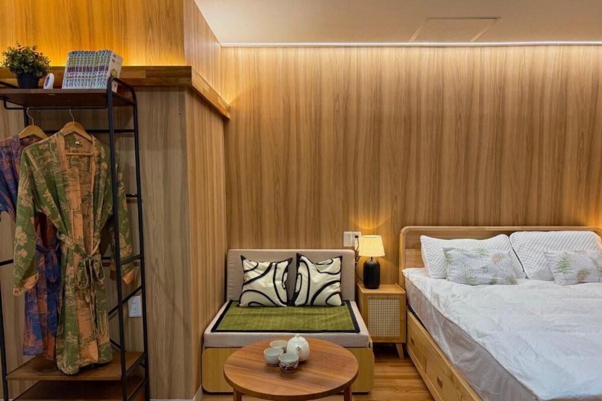 A hotel room with a wooden wall, a bed, a nightstand with a lamp, a small table with a teapot, and a clothing rack with patterned robes.