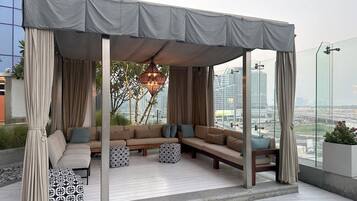 A rooftop lounge with a covered seating area, a patterned ottoman, and a hanging light fixture.