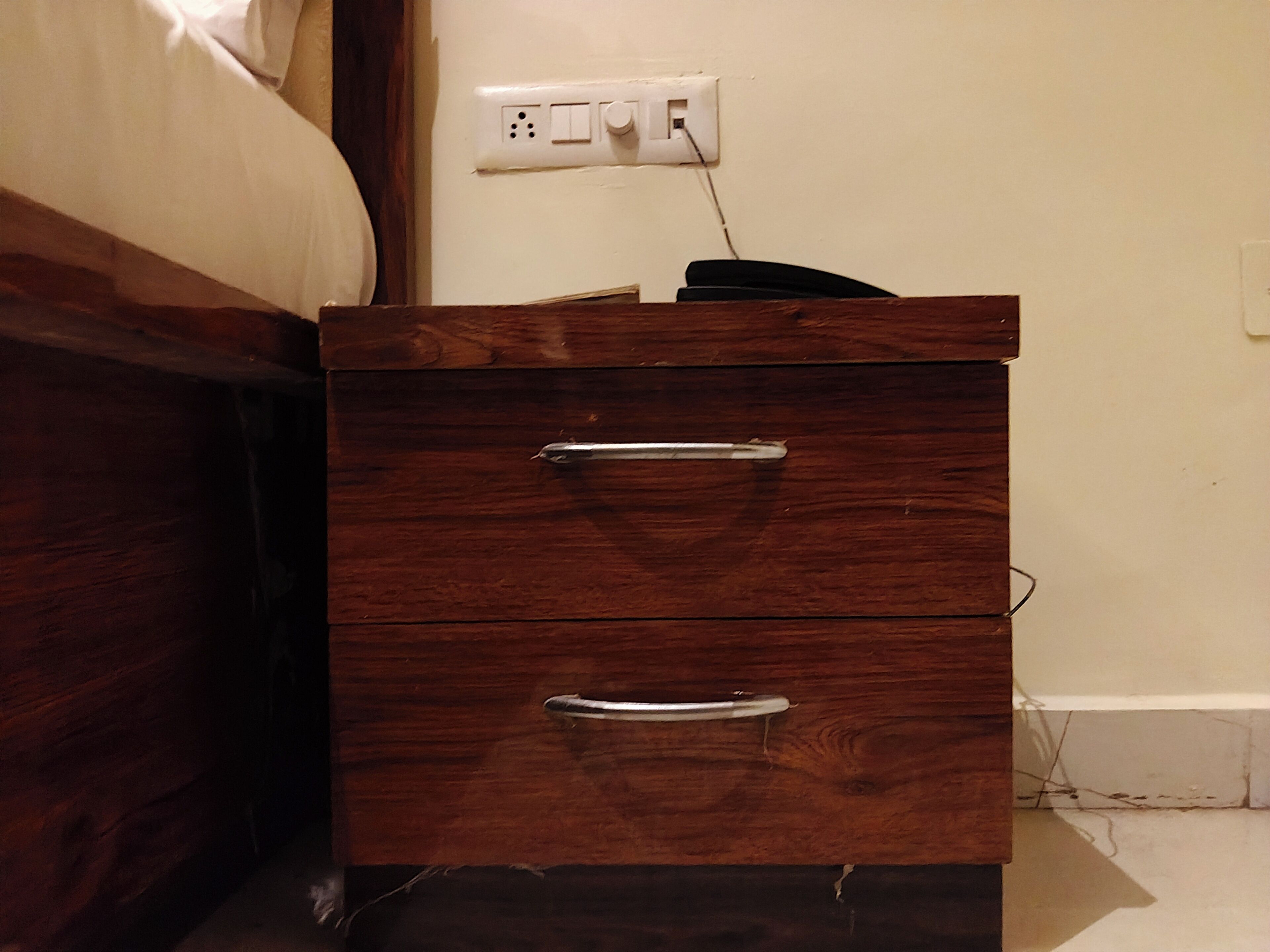 A wooden bedside table with two drawers and a power outlet on the wall.