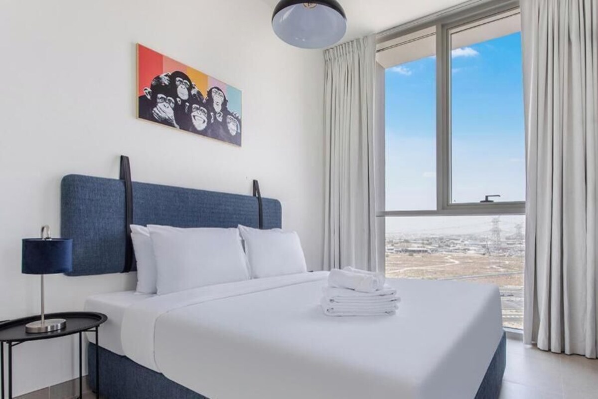 A hotel room with a large bed, a nightstand, a lamp, a painting, and a view of the city through large windows.