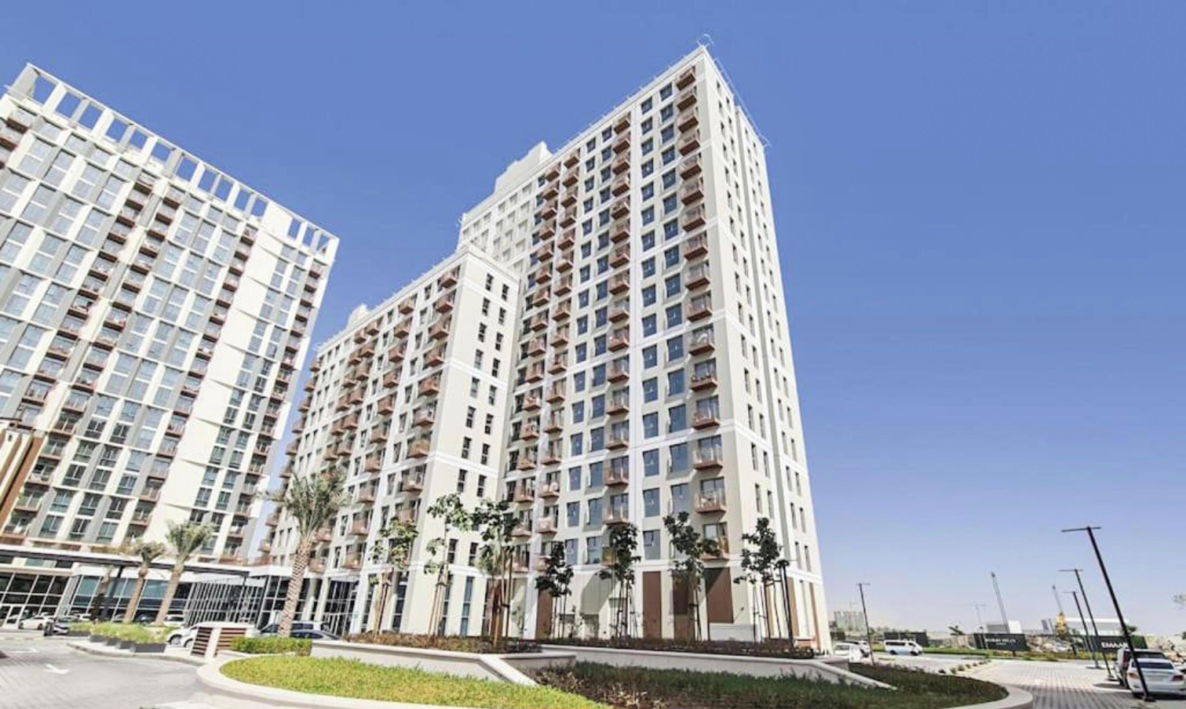 A high-rise residential building with multiple balconies, a landscaped area with palm trees, and a clear sky.