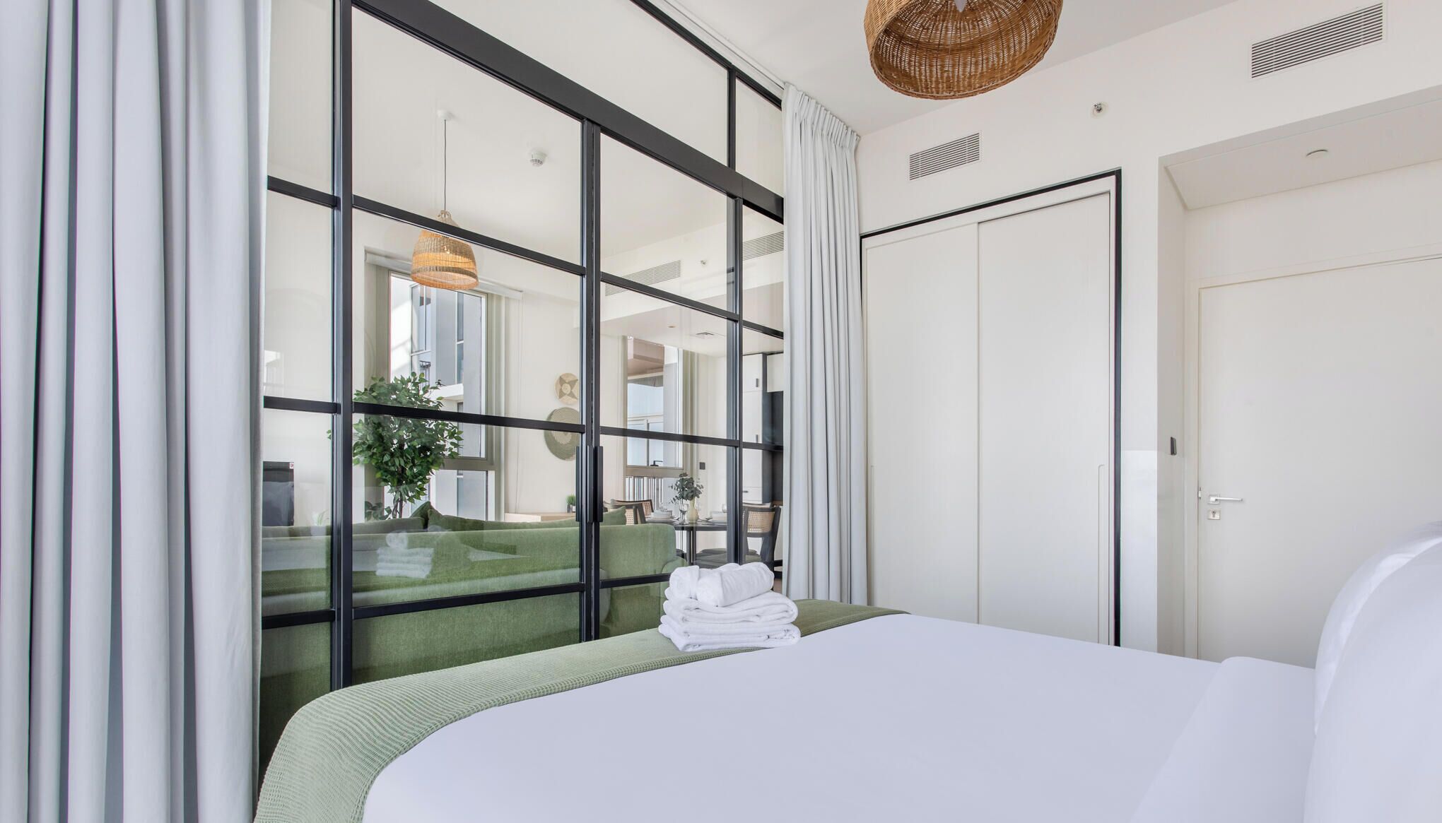 A hotel room with a large bed, a green sofa, a dining table with chairs, a hanging light fixture, and a view through a glass partition.