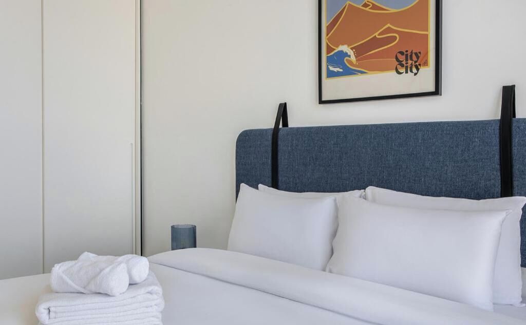 A neatly made bed with white linens and pillows, a blue headboard, a framed picture on the wall, and a stack of white towels on the side.