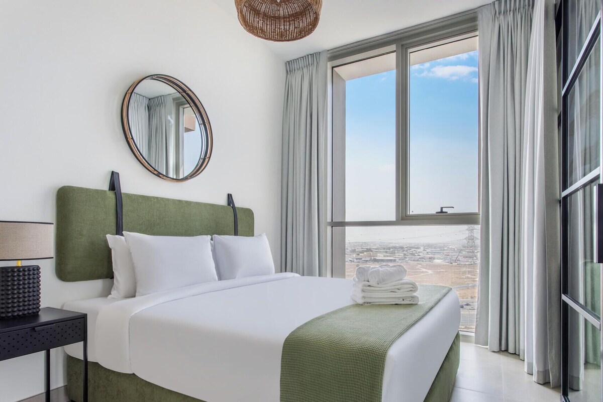 A hotel room with a bed, a nightstand, a lamp, a mirror, and a view of the city through large windows.