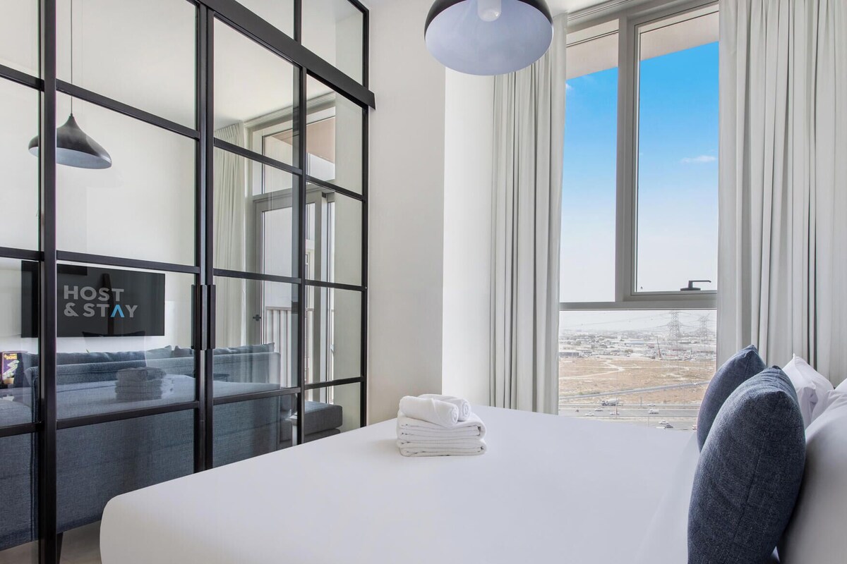 A hotel room with a bed, a window with a view, a sign reading 'HOST & STAY', and a towel on the bed.