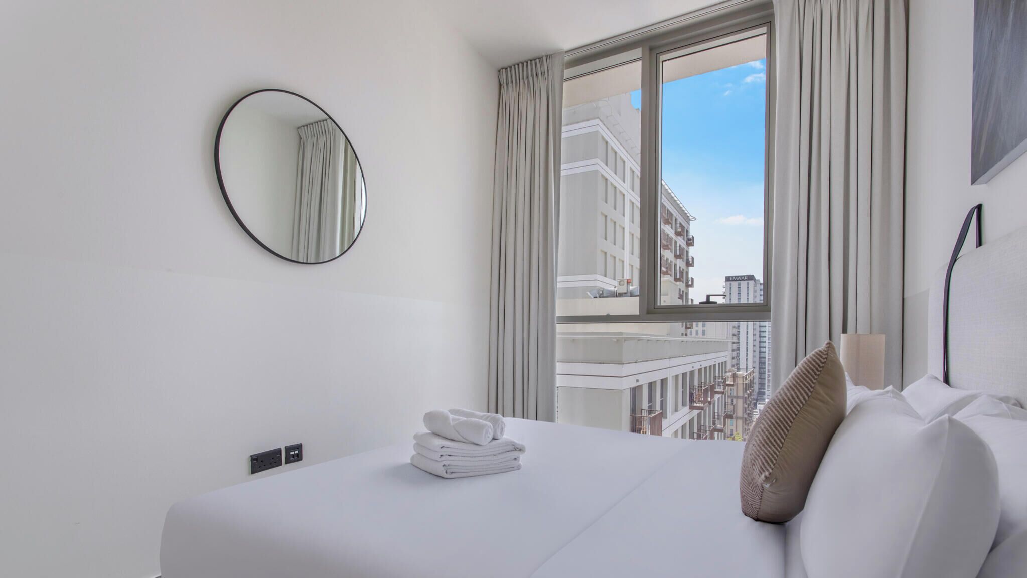 Habitación de hotel con cama, espejo redondo, ventana con cortinas y vistas a edificios.