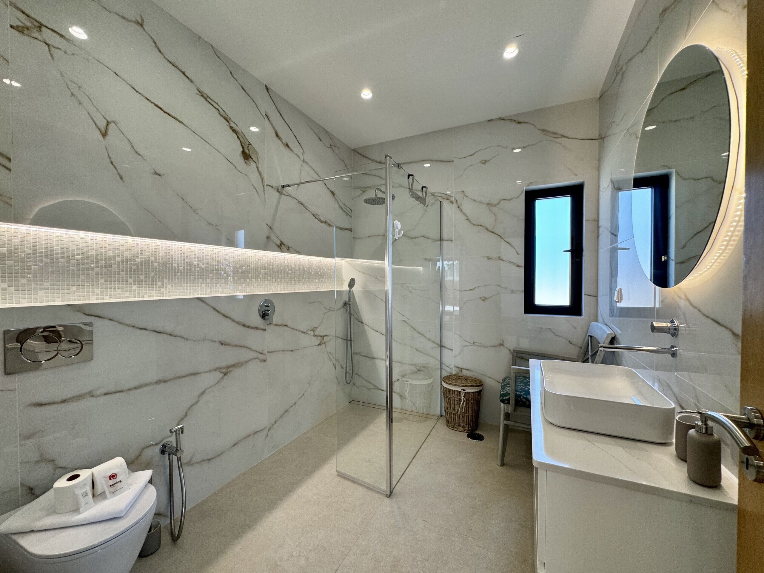 Ein modernes Badezimmer mit einer Glaskabine zur Dusche, einem großen Spiegel und einer Marmorwand.