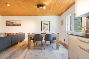 Ein modernes Interieur mit Holzbalkendecke, einer Essecke mit Holztisch und -stühlen, einer Couch und einer Küche mit weißen Schränken und einem Fenster.