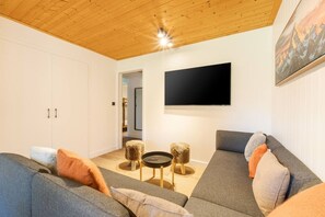 A modern living room with a wooden ceiling, a flat-screen TV mounted on the wall, and a sofa set with cushions. - Géroux- Modern Apt | Quiet | Mountain View | Garden (Chamonix-Mont-Blanc)