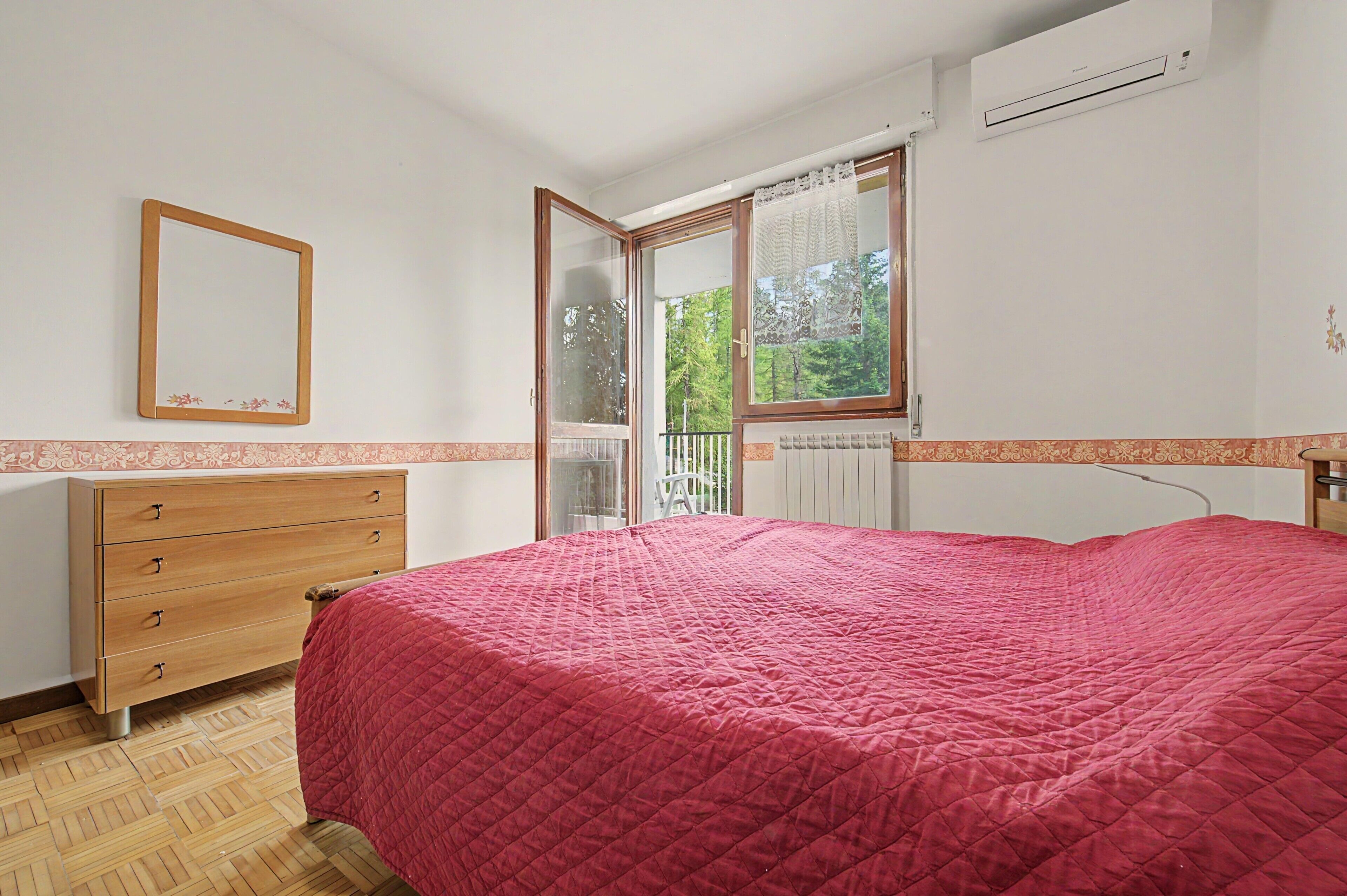 Une chambre à coucher avec un lit, une commode en bois, un miroir et une fenêtre donnant sur des arbres.