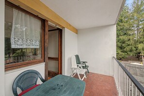 En balkon med et bord og stole, et vindue med blondegardiner og udsigt til træer.