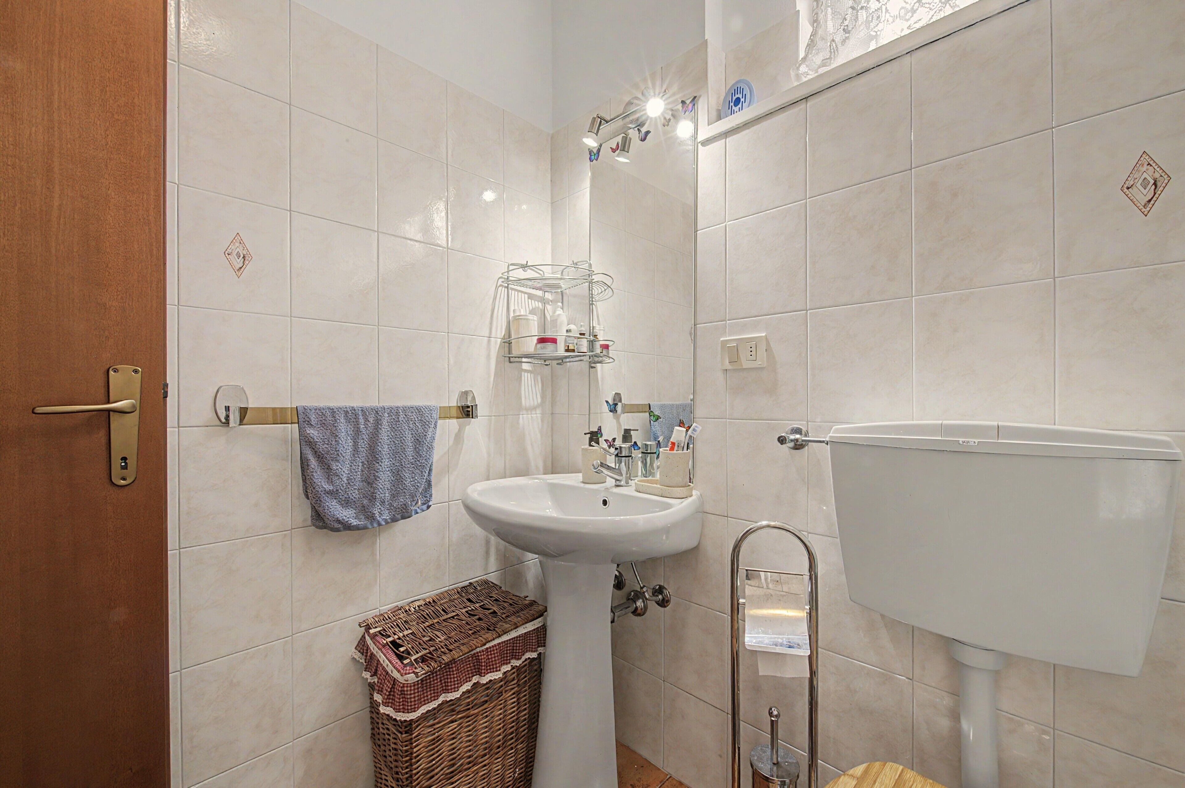 Une salle de bain avec un lavabo blanc, un miroir et des toilettes. On y trouve un porte-serviettes avec une serviette bleue et un panier en osier.