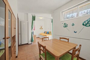 Habitació amb taula de dinar de fusta, cadires, llit amb roba de llit colorida, radiador i finestra amb cortines decoratives.