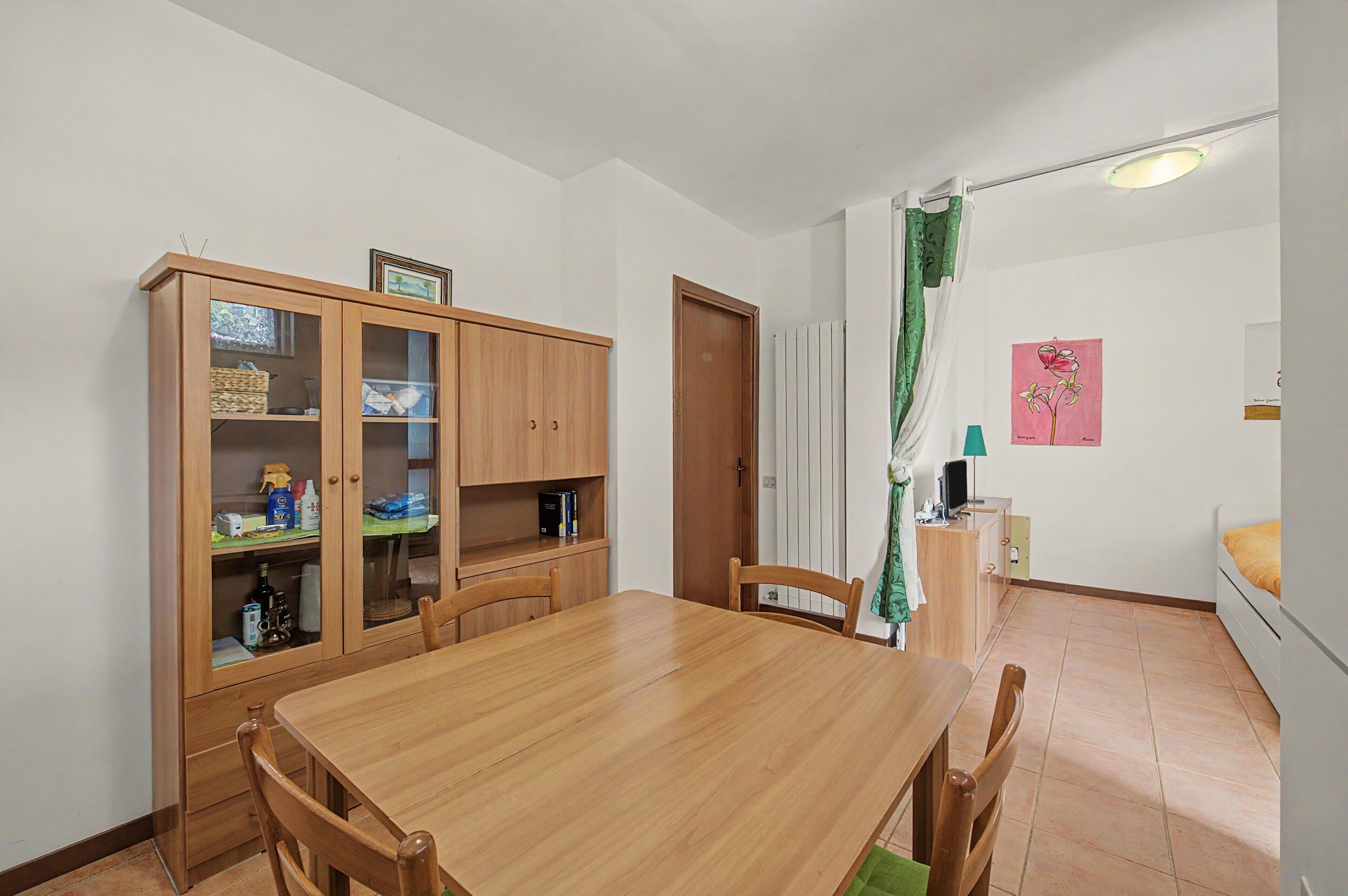 A dining area with a wooden table and chairs, a cabinet with glass doors, a door, a radiator, a green curtain, and a wall with a framed picture.