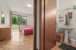 Un dormitori amb sòl de fusta, un llit amb edredó vermell, una finestra que dona a zona verda i un bany amb rentapans i vàter.