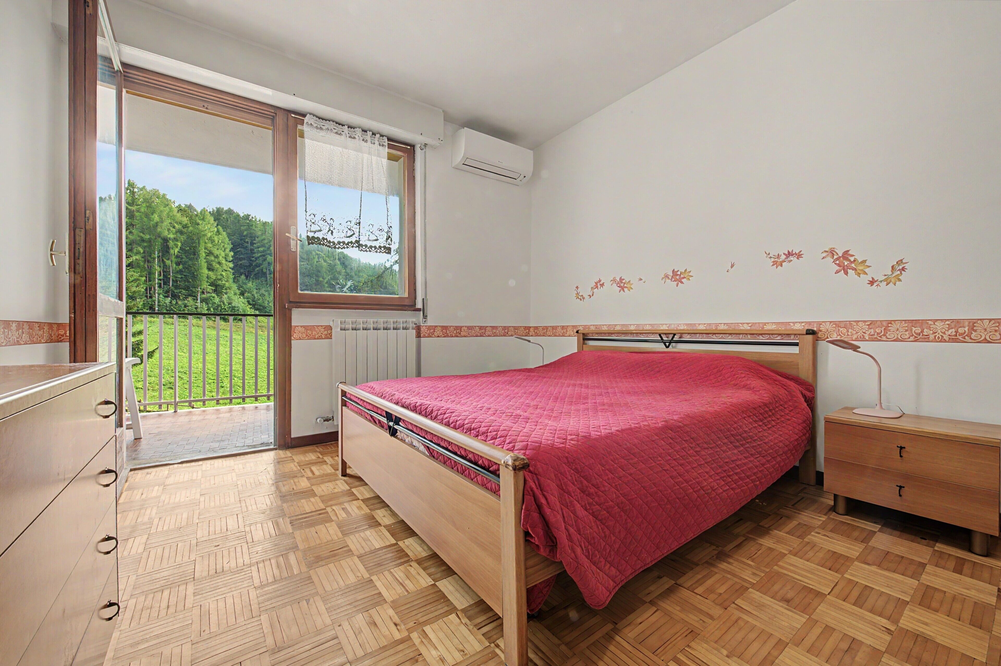 Ein Schlafzimmer mit Bett, Nachttisch, Kommode, einem Fenster mit Blick auf Grünflächen und einer Wand mit Blumenmuster.