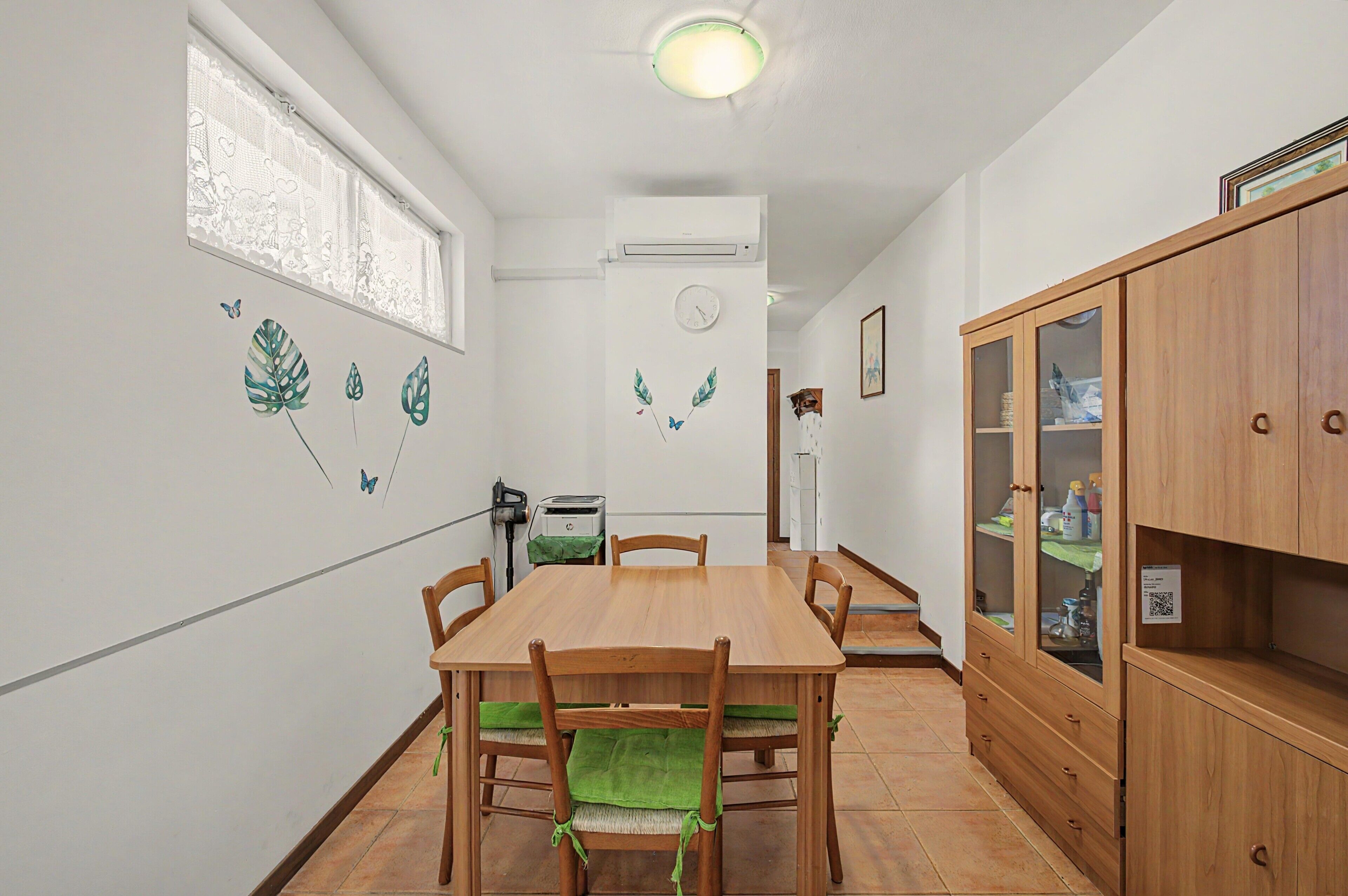 Un coin repas avec une table et des chaises en bois, une fenêtre ornée de rideaux en dentelle, une horloge murale et un meuble avec des portes en verre.
