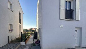 A narrow courtyard with a gravel path, a metal gate, and a small garden with pink flowers. - Small, modern house with a garden, 3 km from the ocean (Aléria)