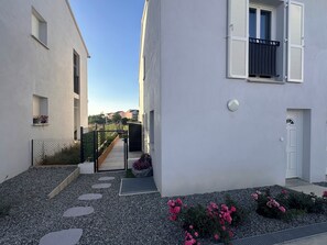 Ein schmaler Innenhof mit Kiesweg, einem Metallgitter und einem kleinen Garten mit rosa Blüten.