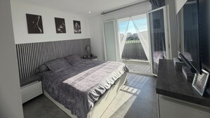 A bedroom with a bed, a nightstand, a TV, and a sliding glass door to a balcony. - Small, modern house with a garden, 3 km from the ocean (Aléria)