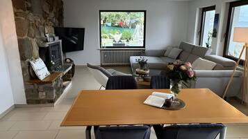 Ein modernes Wohnzimmer mit Kamin aus Stein, einem Esstisch mit Stühlen, einer Couch und einem an der Wand befestigten Fernseher.
