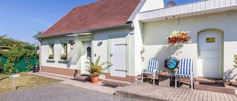 Un edificio con techo inclinado, una puerta adornada con una cesta de flores colgante y un pequeño espacio al aire libre con dos sillas.