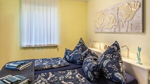Un dormitorio con cama, almohadas, una mesita de noche, una lámpara y una ventana con persianas.