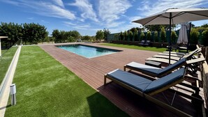 Et rektangulært swimmingpool med træterrasse, omgivet af en velplejet græsplæne og træer. Der er liggestole og et overdækket område med parasol.