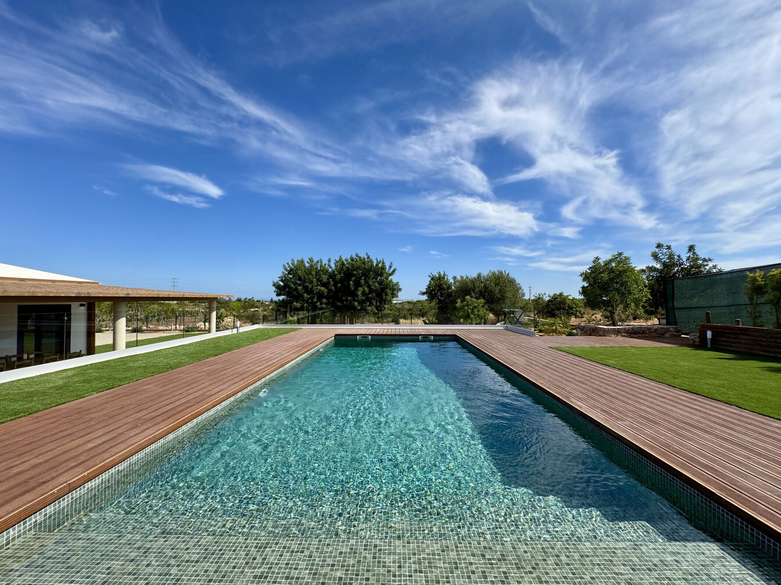 Una piscina rectangular con agua cristalina, rodeada por una terraza de madera y un área verde.