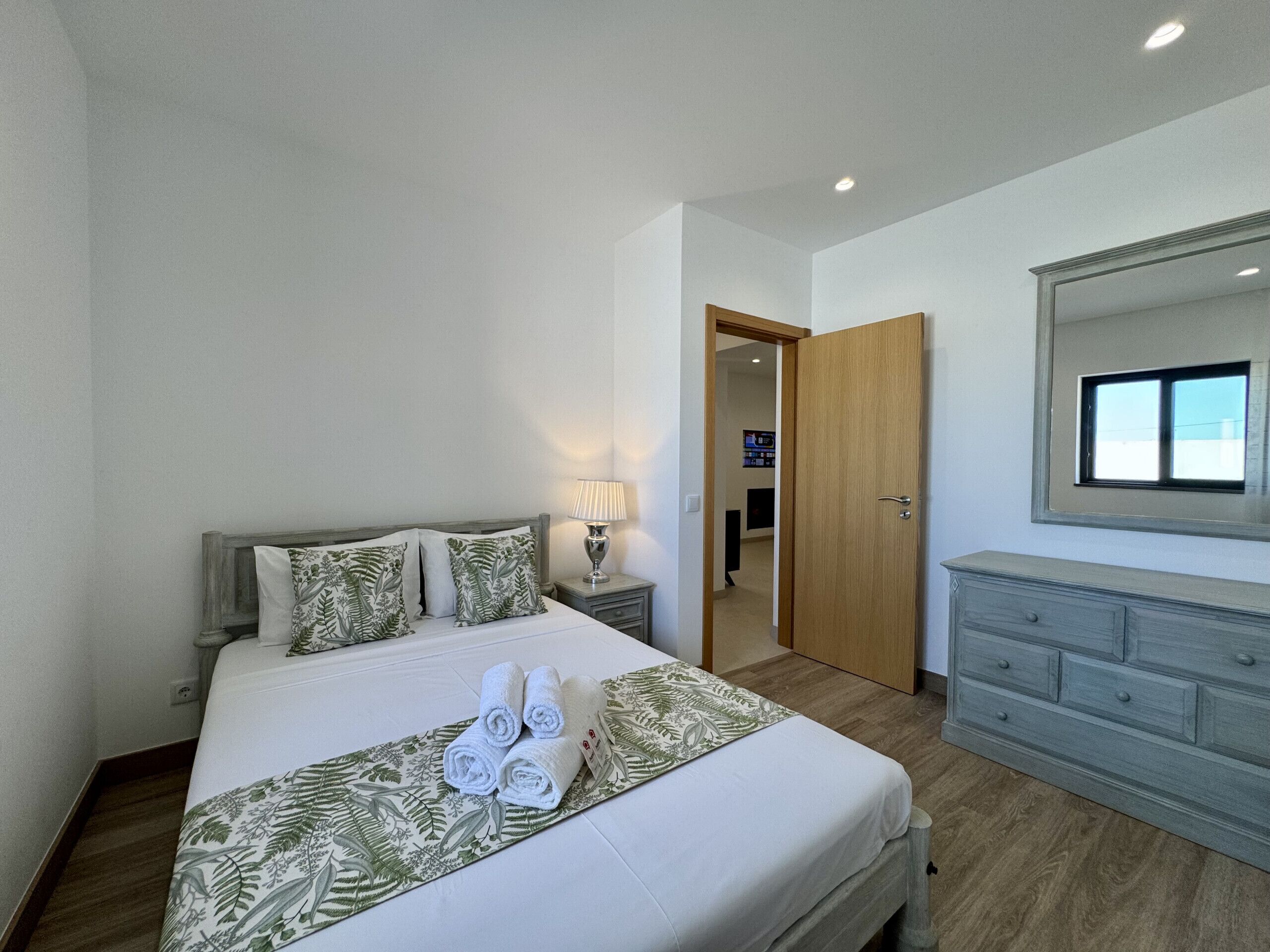 Un dormitorio con cama, mesita de noche, cómoda, espejo y puerta que da a otra habitación.