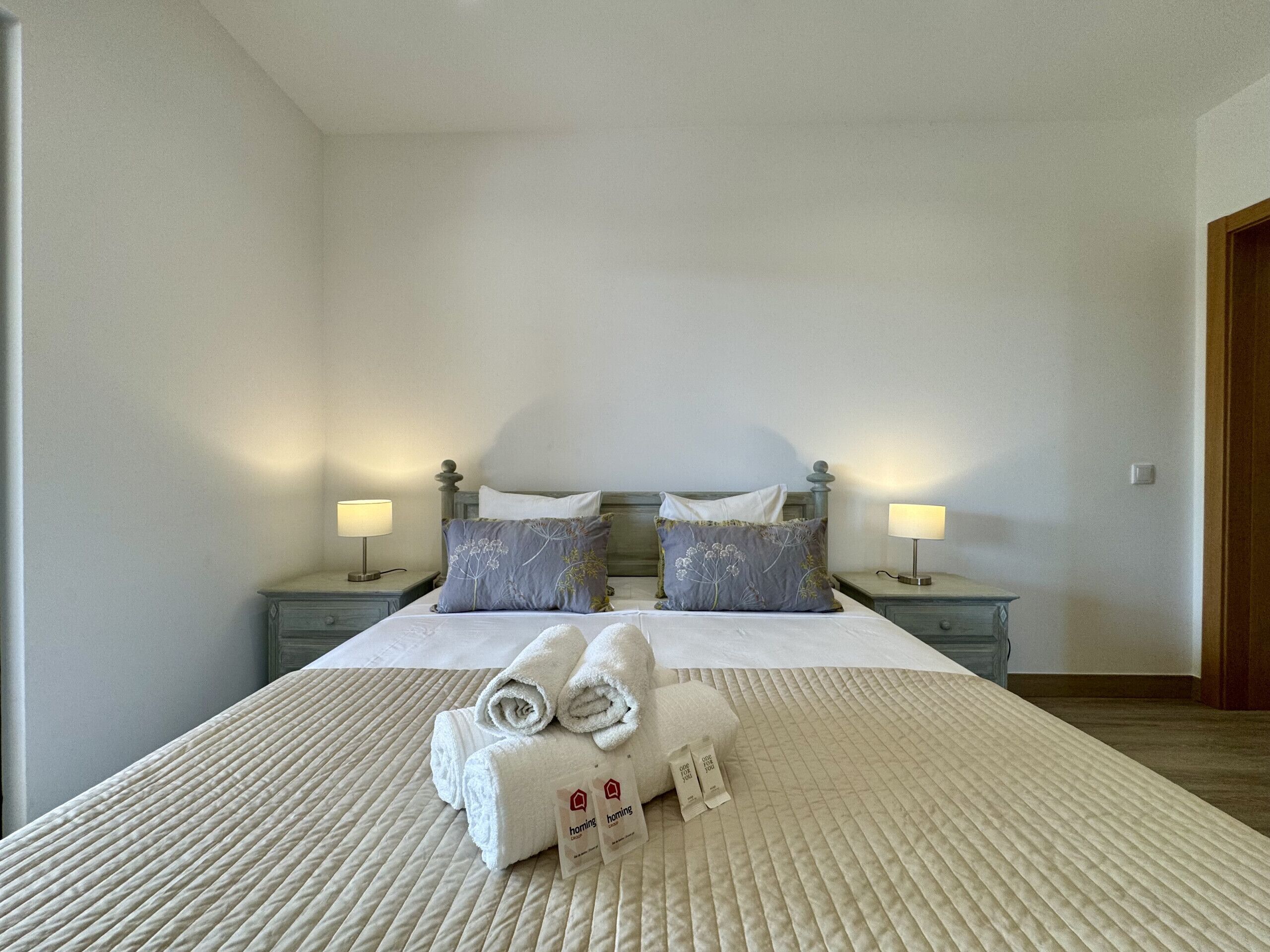 A hotel room with a bed, two bedside tables with lamps, and a neatly made bed with towels and a welcome pack.