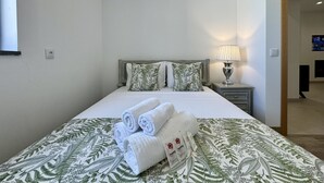 En pænt redt seng med sammenrullede håndklæder, en sengetavlelampe og et blomstret sengetæppe.