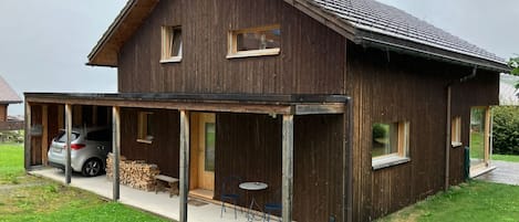 Ett trähus med en inglasad veranda, en bil parkerad utanför och en hög med ved.