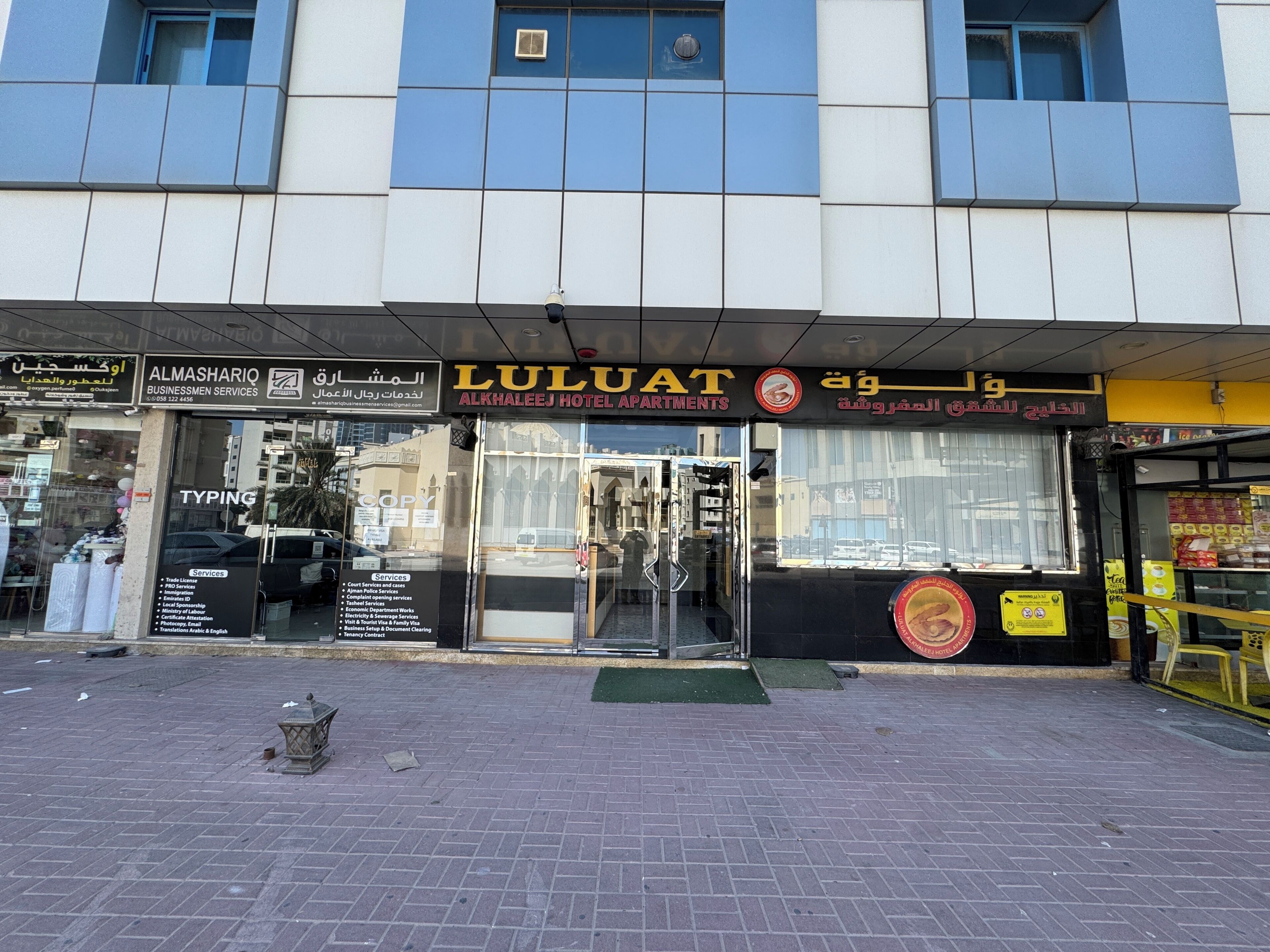 A hotel entrance with a glass door, signage for 'ALMAHLEIJ HOTEL APARTMENTS' and 'LULUAT' services.