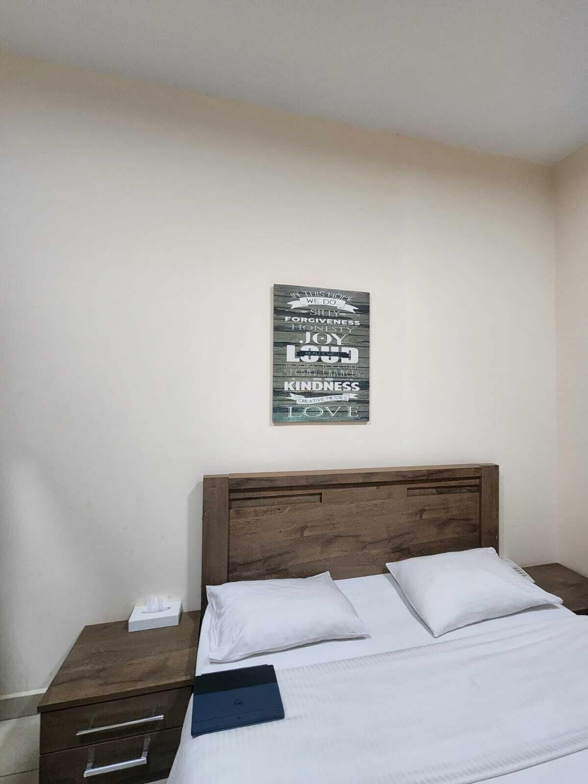 Un llit ben arrenjat amb roba de llenceria blanca, un panell de fusta al capçal i un cartell fixat a la paret amb paraules inspiradores.