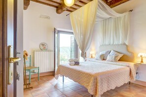 A bedroom with a four-poster bed, a nightstand with a lamp, a chair, and a view of the outdoors.