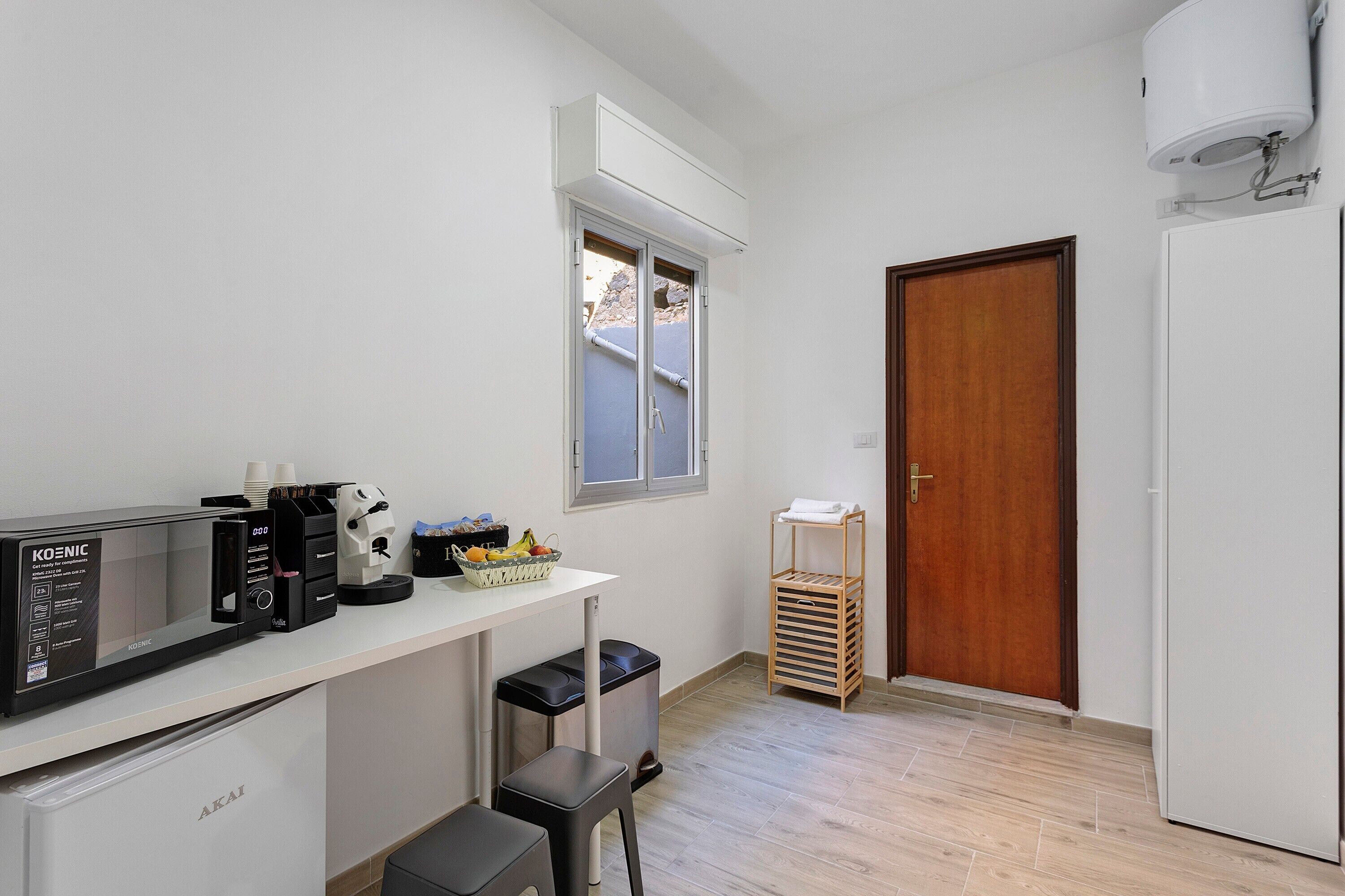 Eine kompakte Küche mit Mikrowelle, Kaffeemaschine und einem kleinen Kühlschrank. Über der Arbeitsplatte befindet sich ein Fenster und rechts eine Tür.