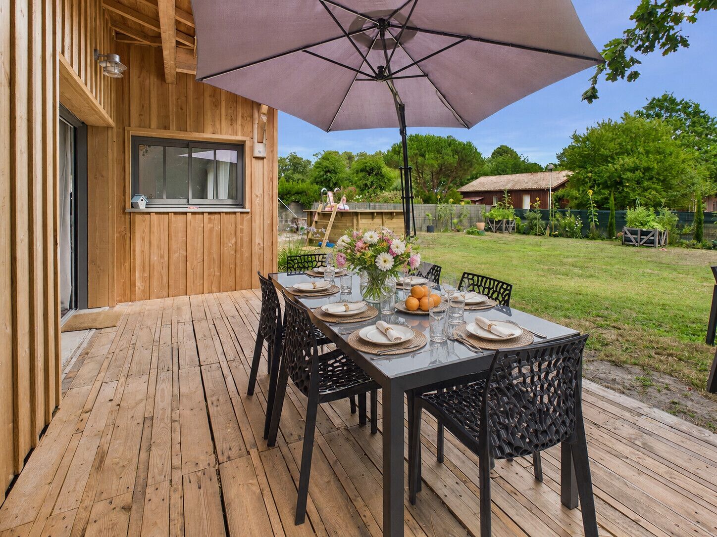 An outdoor wooden deck with a table set for dining, surrounded by chairs, and an umbrella providing shade.