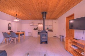 A living room with a fireplace, a dining area with chairs, a TV on a wooden stand, and a kitchen in the background. - Heimeliger Doppelbungalow mit Terrasse im Ferienpark Mirow (Granzow)
