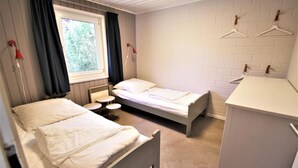 Ein Zimmer mit zwei Betten, einem kleinen Tisch, einem Fenster mit Vorhängen, einem Heizkörper und einem weißen Schrank.