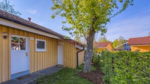 En etagers træbygning med dør og vindue i hvidt, et lille træ foran og en belagt sti.