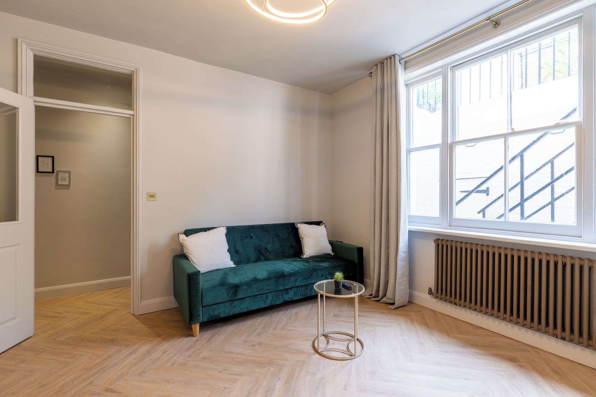 Una habitación con un sofá verde, una mesita redonda con una planta, un radiador y una ventana que da a una escalera.