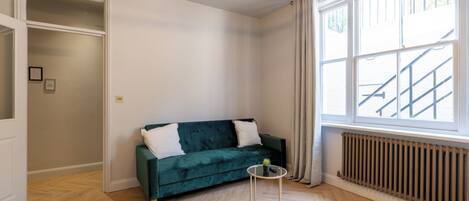 Una habitación con un sofá verde, una mesita redonda con una planta, un radiador y una ventana que da a una escalera.