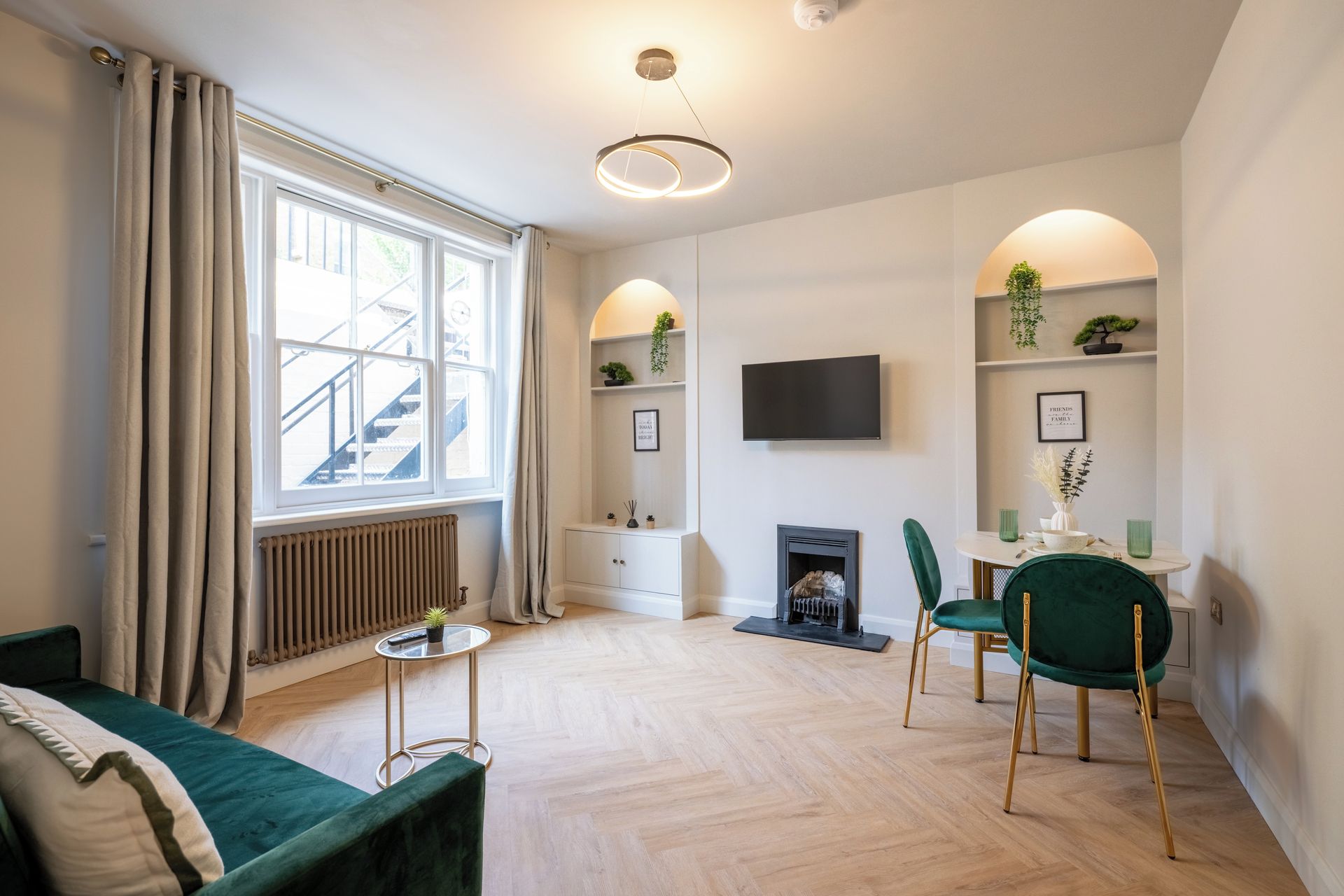 A modern living room with a fireplace, a dining area with green chairs, a TV mounted on the wall, and a staircase leading to an upper floor.