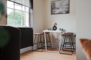 Ein Zimmer mit Fenster, einem Tisch mit Hocker, einem Heizkörper und einem gerahmten Bild an der Wand.