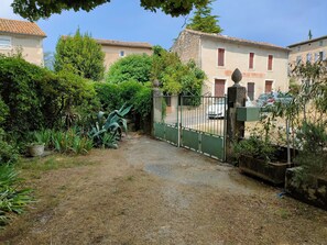 A gravel path leading to a gated entrance with a fence, surrounded by greenery and buildings with tiled roofs. - Large Country House (Brouzet-lès-Alès)