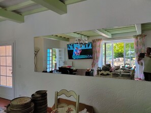 A room with a television mounted on the wall, a window with curtains, a table with stacked plates, and a chair. - Large Country House (Brouzet-lès-Alès)