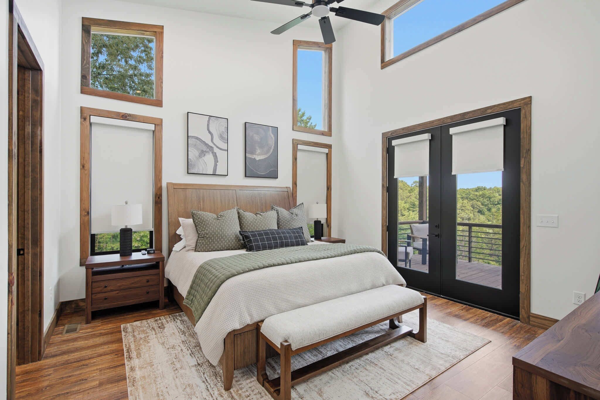 A bedroom with a bed, nightstands, a bench, a ceiling fan, and large windows.